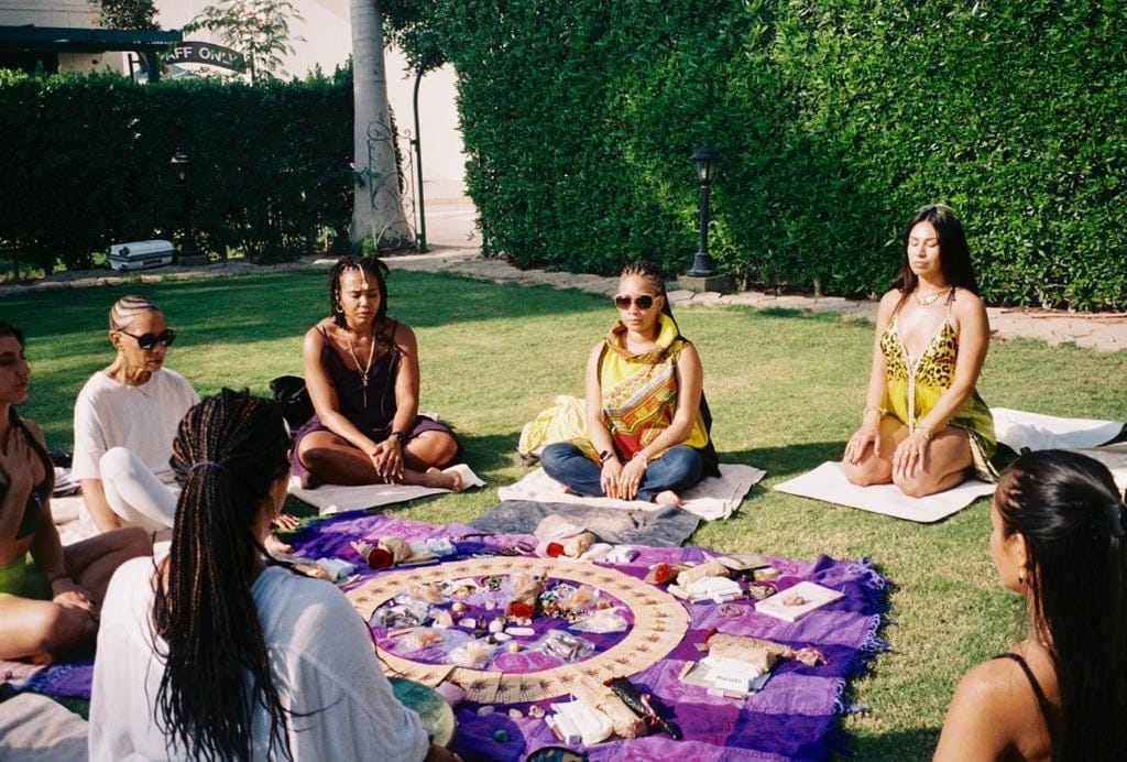 Women in a circle on the grass around a purple blanket with flowers, cards, and ceremonial objects