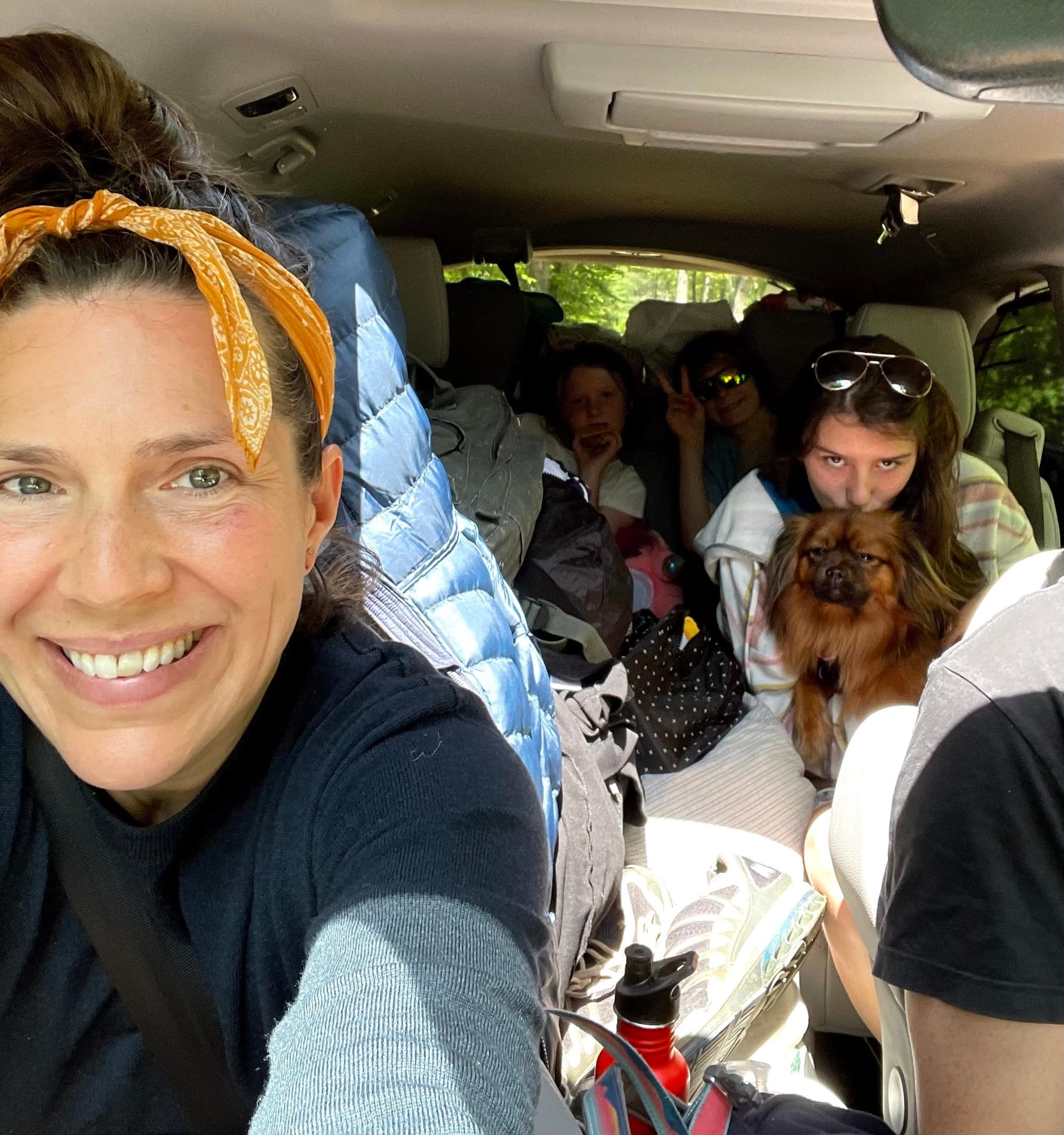 Woman, hair tied up in yellow bandana in a van packed with kids, clothes and a brown dog.