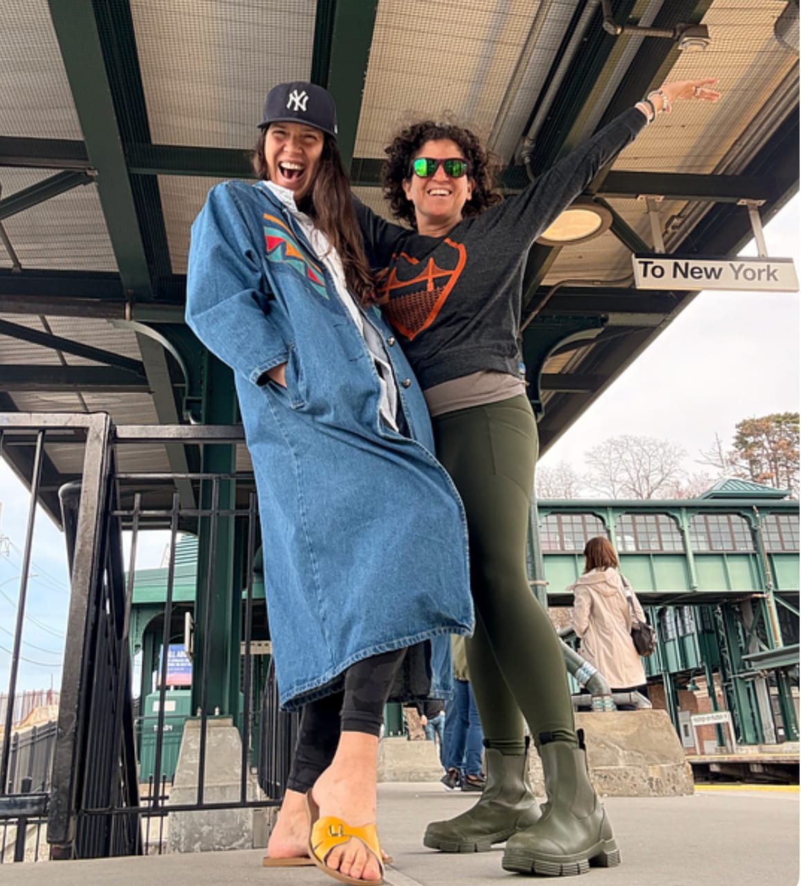 Two joyful white women with brown hair, one in long jean jacket, the other in green tights and rubber boots.