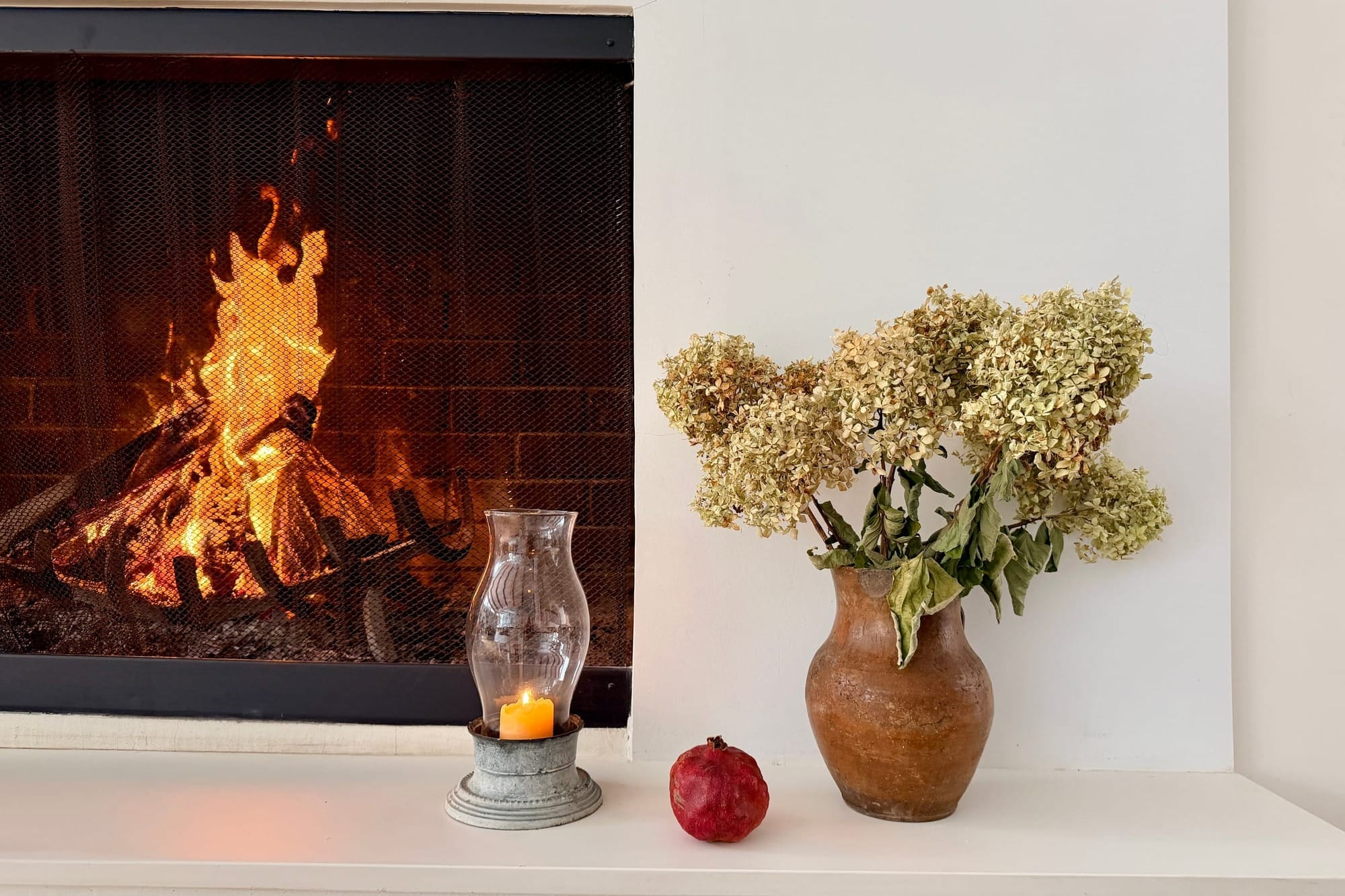 A fire crackles beside a candle, pomegranate, and clay vase of dried hydrangeas