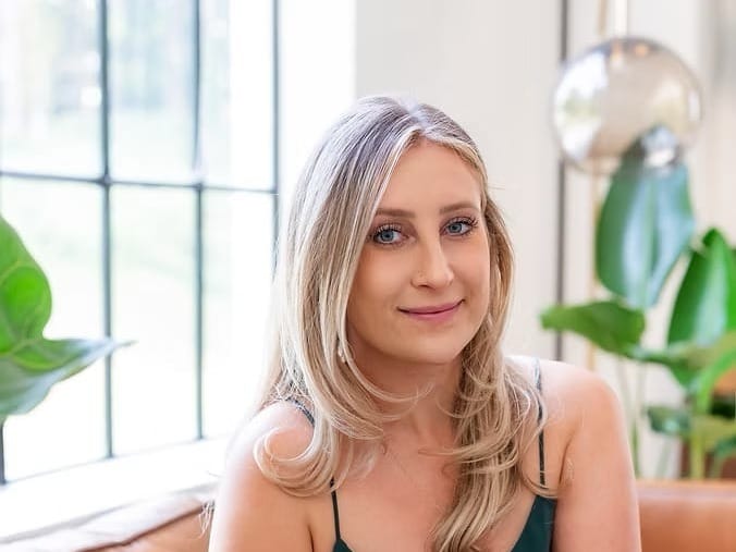 Woman with long blond hair and painted fingernails sits on a leather couch in light-filled studio