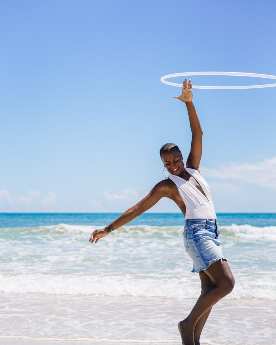Black woman with short natural hair, hoop earrings, and a short denim skirt and white tank top twirls a hula hoop over her head on the ocean's edge 