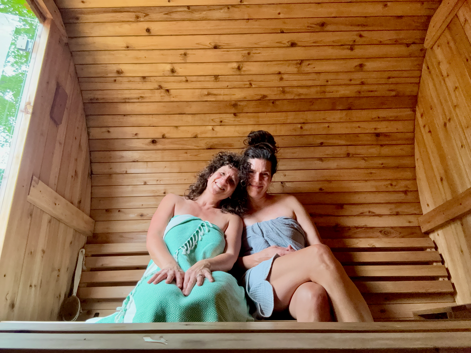 Two white women with brown hair sit in a cedar barrel sauna wrapped in green and grey towels
