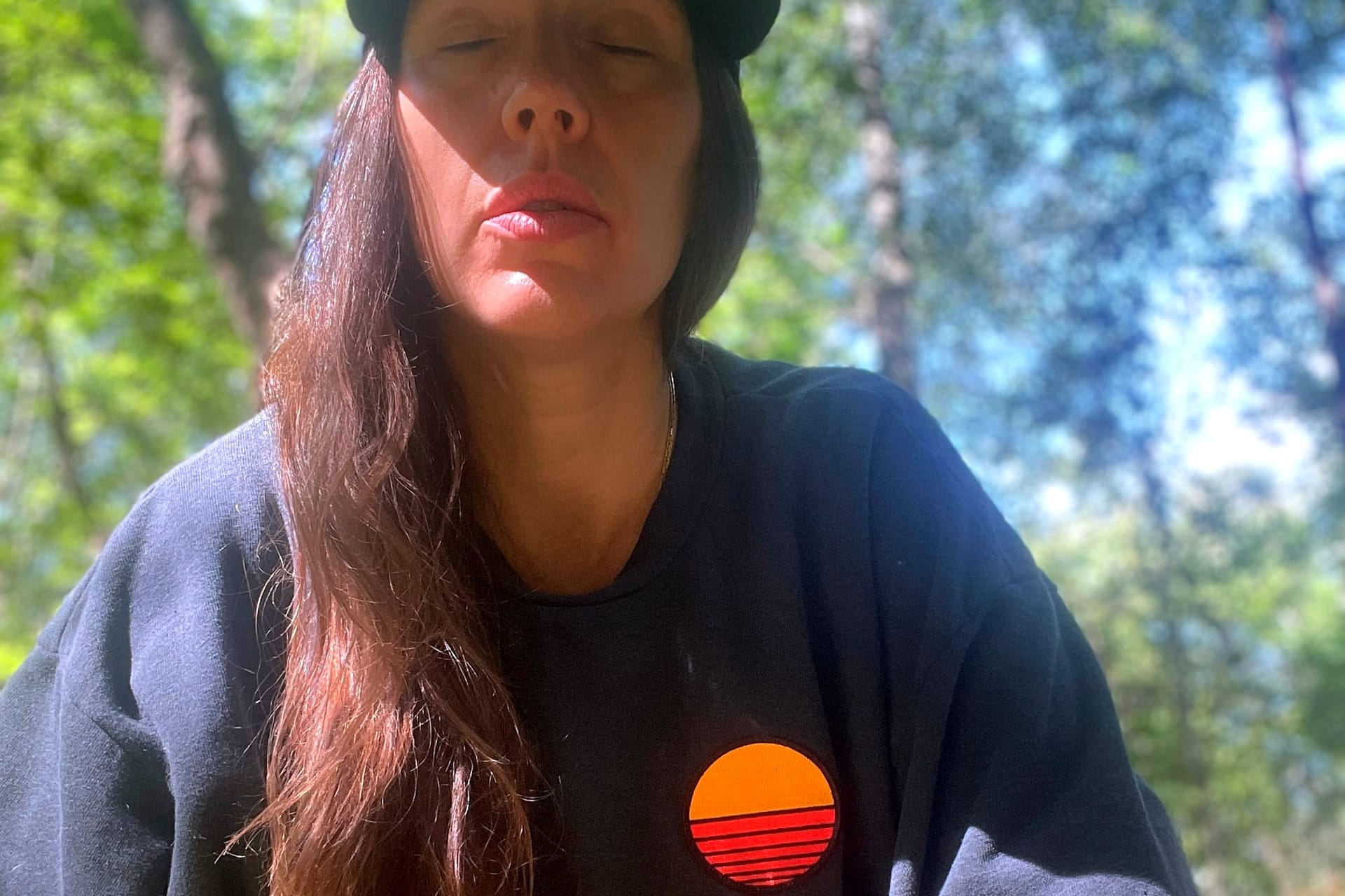 Sitting cross-legged in the woods, a white woman in a navy sweatshirt exhales deeply with eyes closed. 