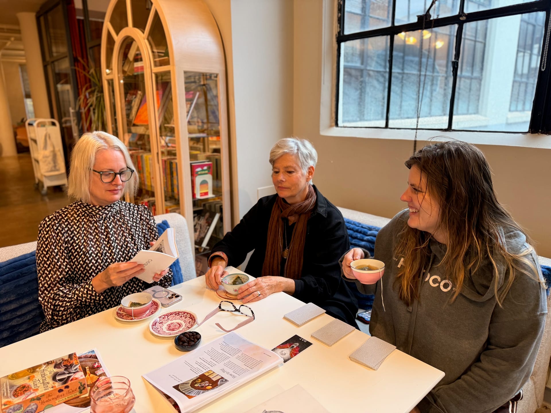 Three women hold teacups and read oracle cards in a cozy professional office space