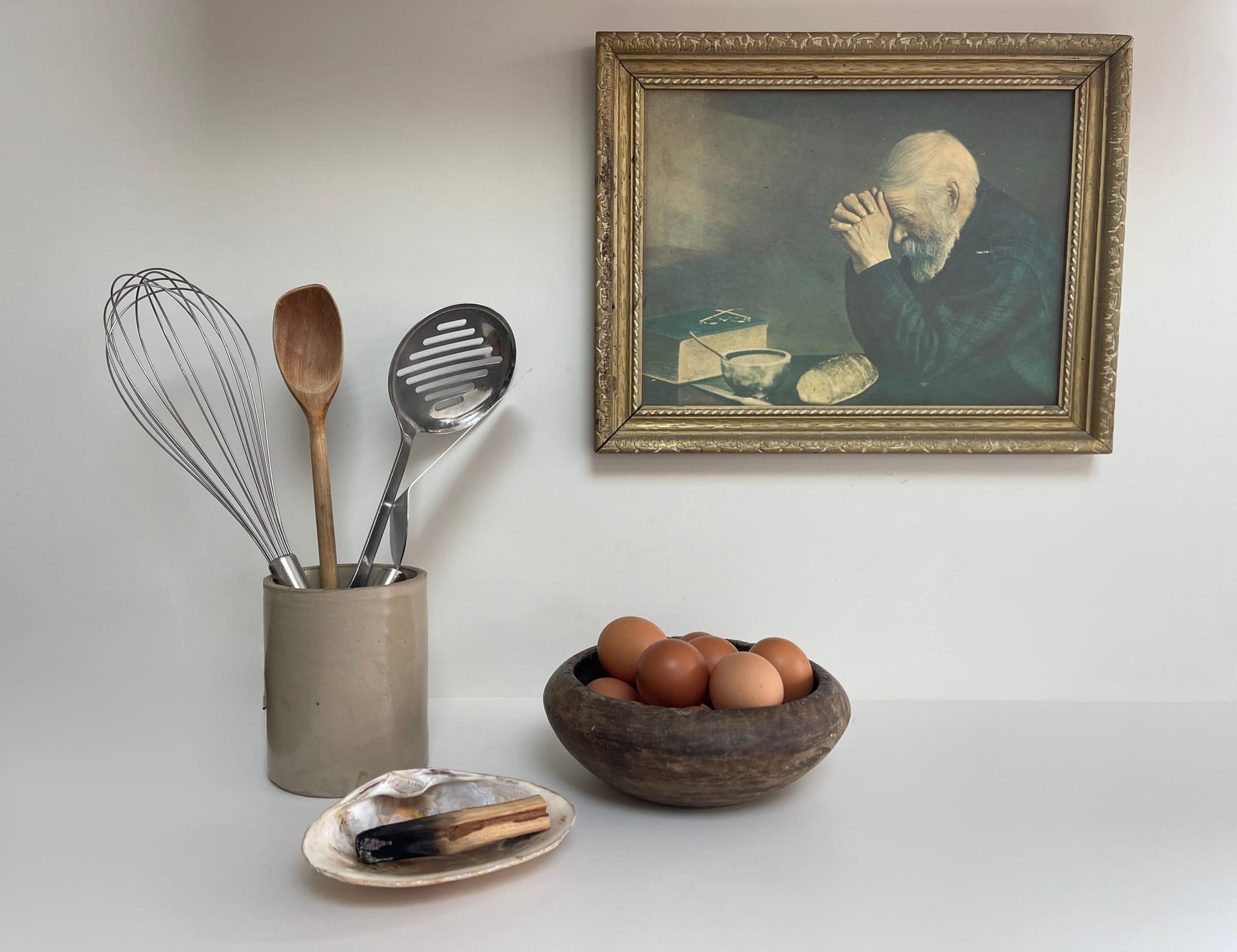 Cooking utensils sit in a crock pot, palo santo nestled in a shell in front of a wooden bowl full of brown eggs all on the counter in front of a painting of an old man praying over his bread