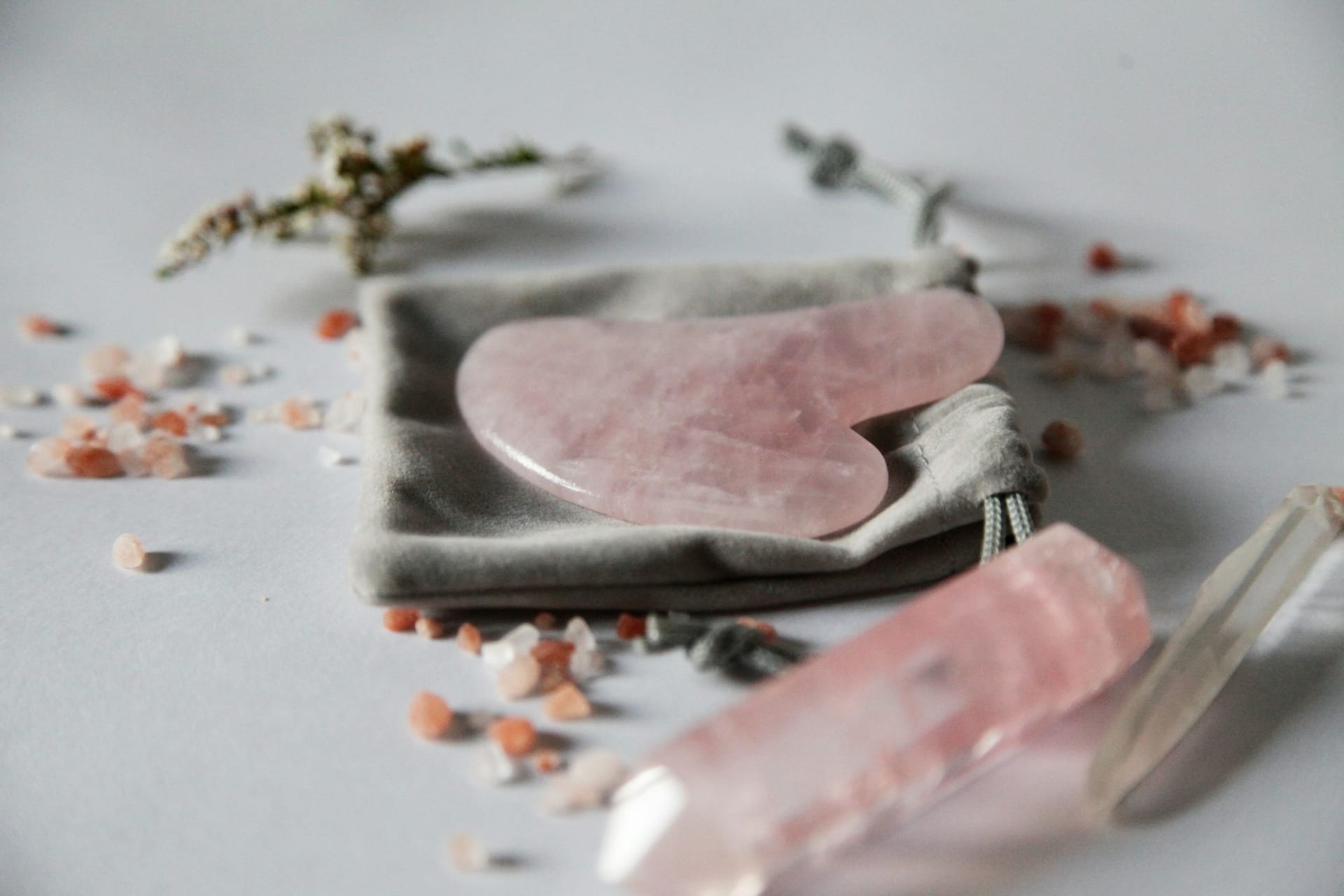 Pink gua sha tool in the shape of a heart in rose quartz with crystal in the foreground on grey velvet bag with pink Himalayan salt spread around