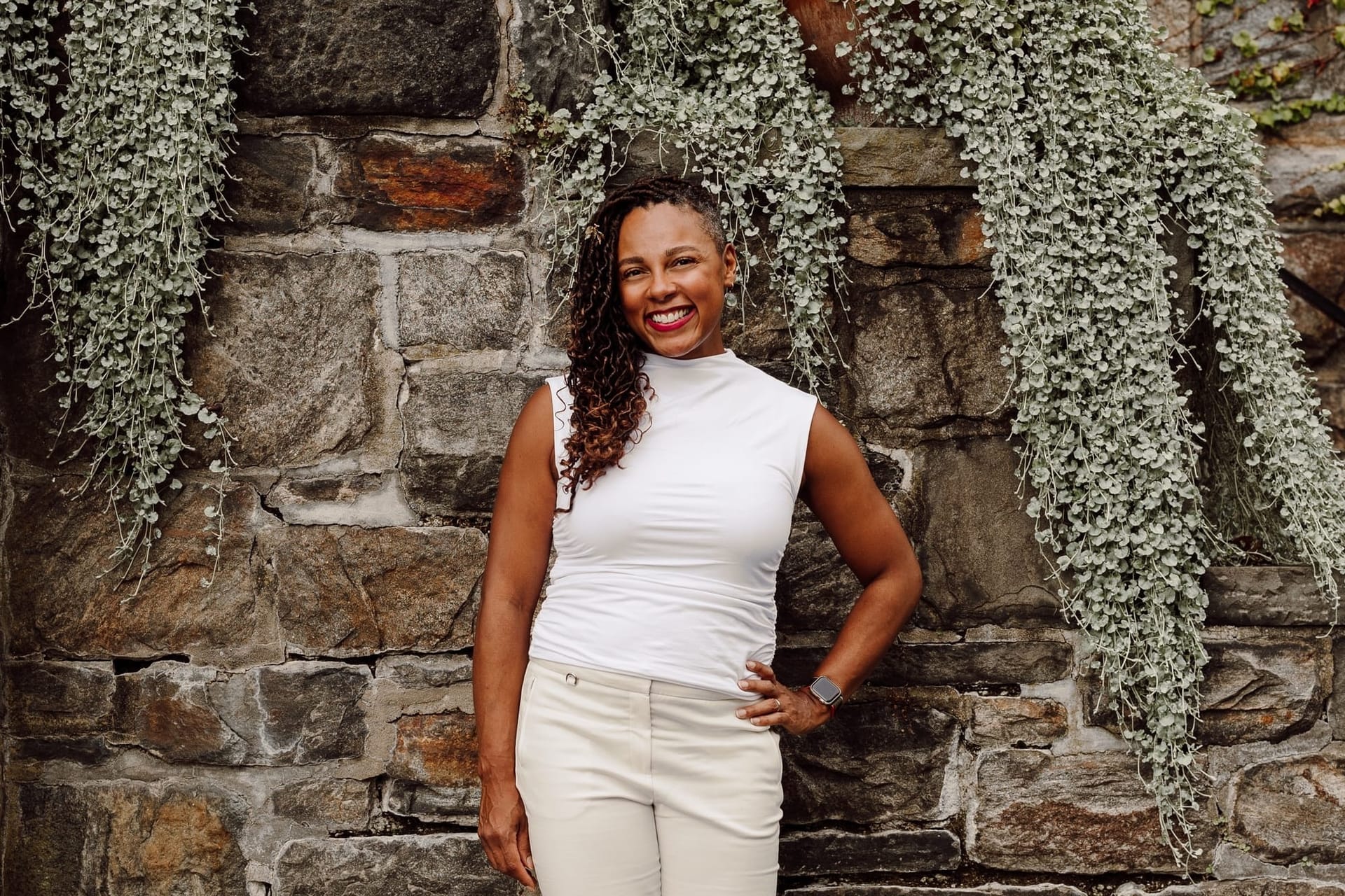 Woman with long braids smiles wearing all white in front of a vine-covered stone wall