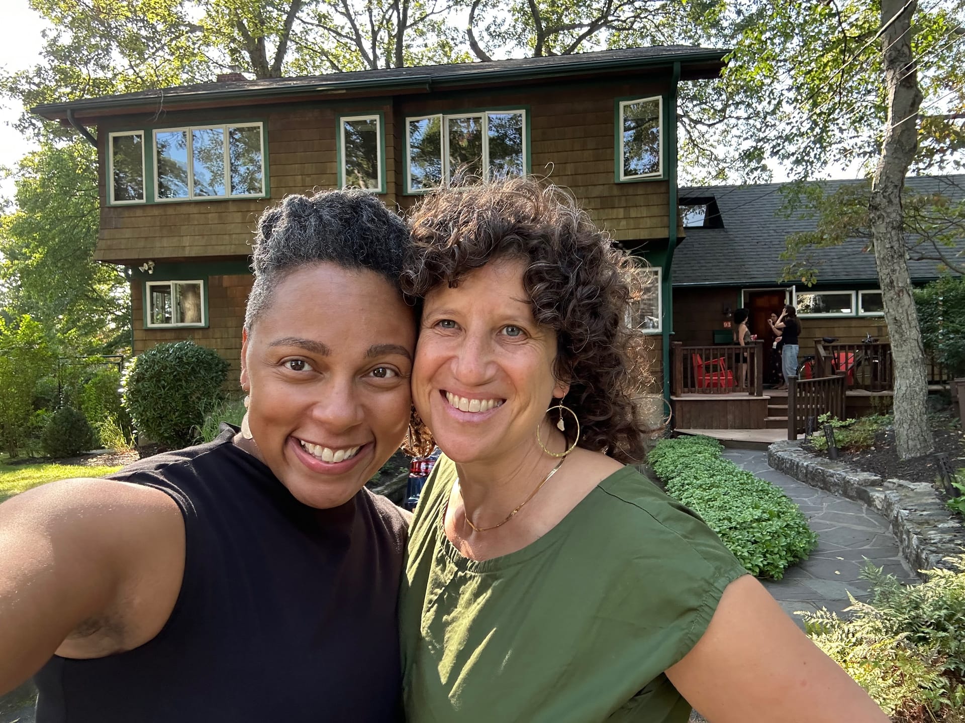 Two women take a selfie on a sunny day in front of a wooden house with tall oaks