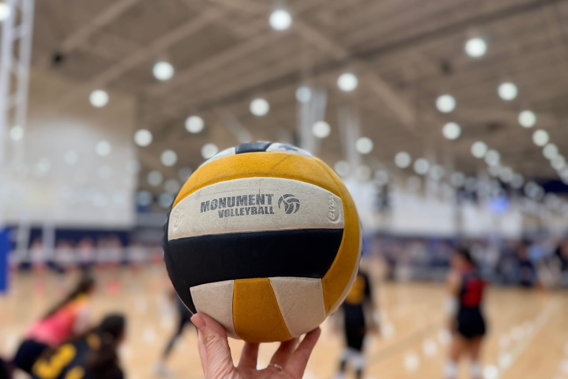 A woman's hand holds a volleyball against the backdrop of a brightly lit gym