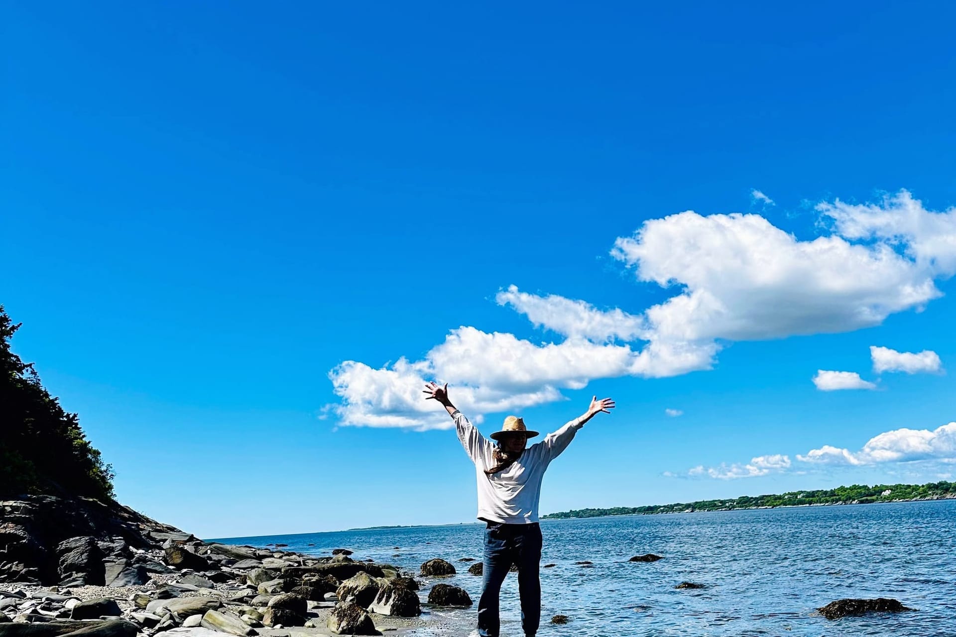 A woman stands in blue pants and a wide brim hat with arms outstretched taking in the ocean