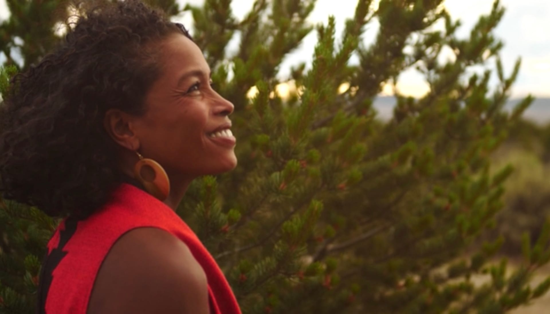A black woman in a red top with large gold hoop earrings stands smiling in front of an evergreen bush